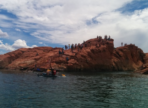 A group of people on a rocky shoreline, with kayakers in the water and a cloudy sky above.