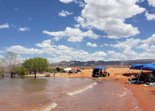 A sandy beach by a lake with tents and people enjoying the sunny day under a blue sky with fluffy clouds.