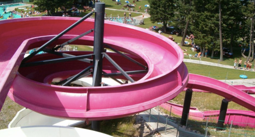A pink water slide spirals down into a pool area surrounded by trees and sunbathers enjoying a summer day.