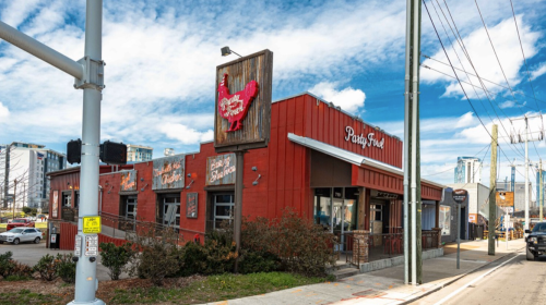 A red restaurant building with a rooster sign, surrounded by greenery and power lines, under a blue sky.