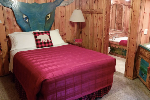 Cozy bedroom with a red quilt, wooden walls, a bull mural, and a bear-themed pillow, featuring an adjoining bathroom.