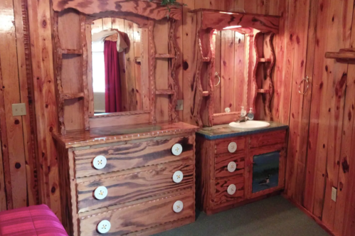 Wooden vanity and dresser with unique designs, featuring a sink and illuminated mirrors in a rustic room.