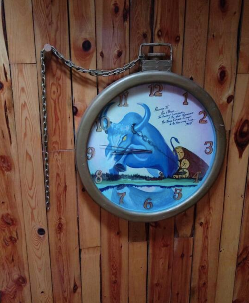 A large, round wall clock with a blue bull design, hanging on a wooden wall, featuring a chain and decorative numbers.