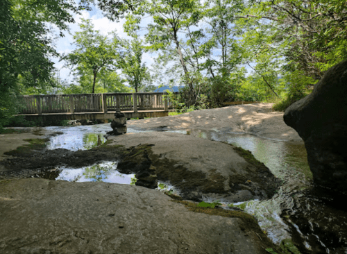A serene landscape featuring a stream, smooth rocks, and a wooden bridge surrounded by lush greenery.