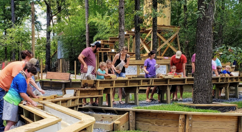 Families and children enjoy a hands-on activity at an outdoor water and sand play area surrounded by trees.