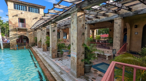 A rustic building with a poolside view, featuring columns, greenery, and a sunny sky.