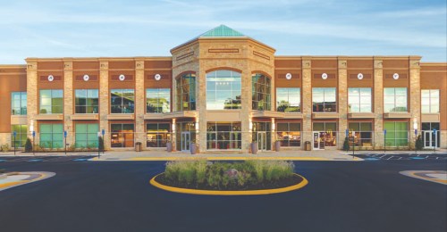 Modern retail building with large glass windows, stone accents, and a landscaped entrance. Clear blue sky above.