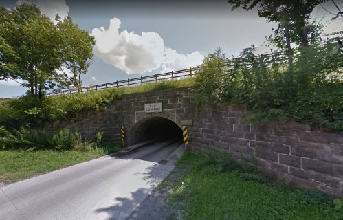A stone tunnel entrance surrounded by greenery, with a road leading through and a sign above indicating clearance height.