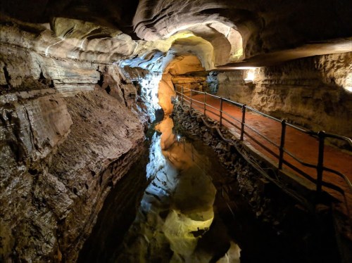 A dimly lit cave with a narrow walkway beside a calm, reflective water surface. Rock formations line the walls.