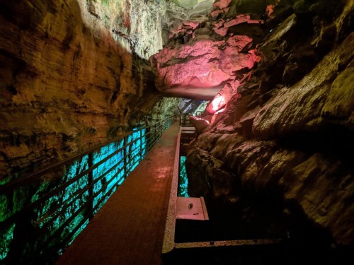 A narrow walkway through a colorful, illuminated cave with rocky walls and water below.