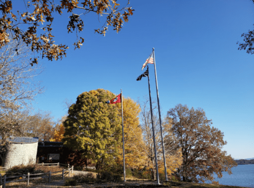 Three flags stand tall near a lake, surrounded by colorful autumn trees under a clear blue sky.