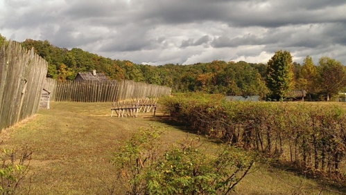 A rustic landscape featuring a wooden fence, a few small buildings, and a backdrop of trees under a cloudy sky.