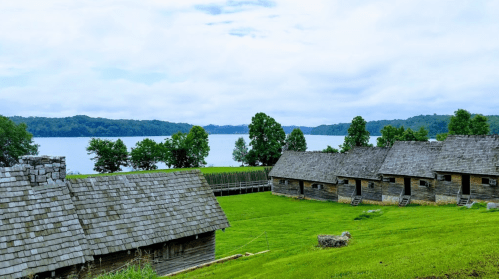 A scenic view of wooden cabins by a lake, surrounded by green grass and trees under a cloudy sky.