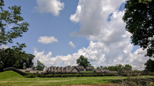 A historic village with wooden structures under a blue sky filled with fluffy clouds and green grass in the foreground.