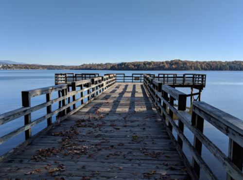 A wooden pier extends into a calm lake, surrounded by trees with autumn foliage under a clear blue sky.