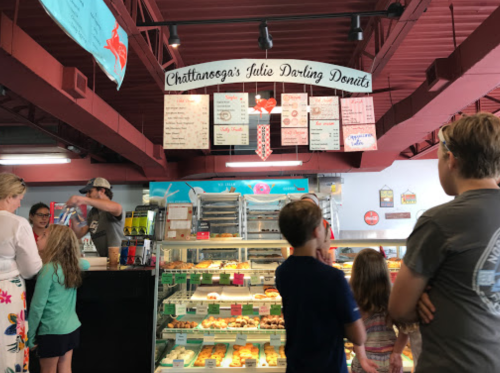 A busy donut shop with a menu overhead, customers waiting in line, and a display case filled with colorful donuts.