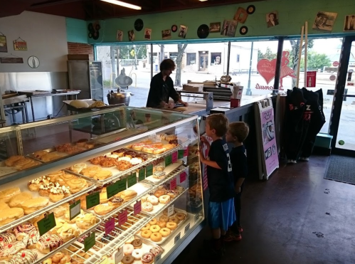 Two children gaze at a display case filled with pastries in a bakery, while an employee assists a customer in the background.