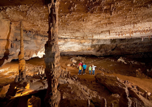 A group of people exploring a large cave with stalactites and stalagmites, illuminated by soft lighting.