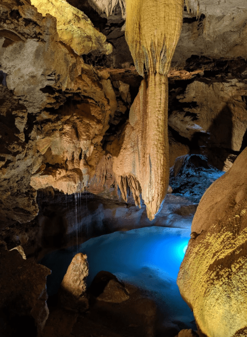 A cave interior featuring stalactites, rock formations, and a blue-lit pool of water below.