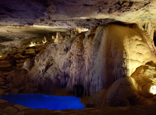 A dimly lit cave with stalactites and stalagmites, featuring a small blue pool of water illuminated by soft lights.
