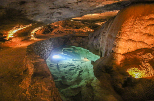A dimly lit cave interior with illuminated rock formations and a clear pool of water reflecting the light.