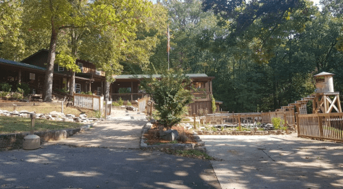 A scenic view of a rustic lodge surrounded by trees, featuring a pathway, gardens, and a wooden deck.