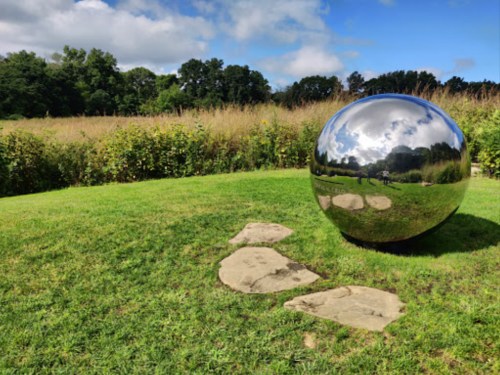 A reflective metal sphere sits on grass, surrounded by a field and trees under a partly cloudy sky.