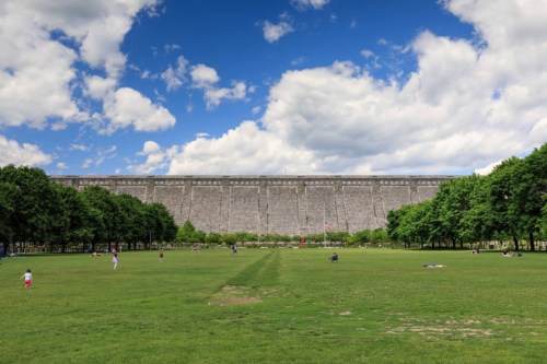 A wide grassy area with people enjoying the outdoors, framed by trees and a large stone wall under a blue sky with clouds.