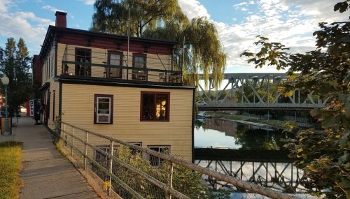 A two-story building by a river, with a bridge in the background and trees reflecting in the water.
