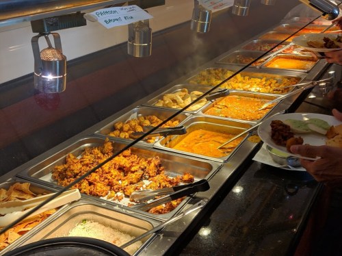 A buffet with various dishes in metal trays, including curries, fried items, and rice, with steam rising.