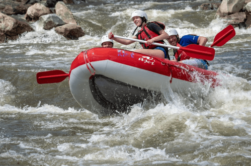 Three people in a red raft navigate whitewater rapids, smiling and paddling through the turbulent water.