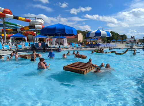 A busy water park pool with people swimming, playing, and enjoying water slides under a sunny sky.