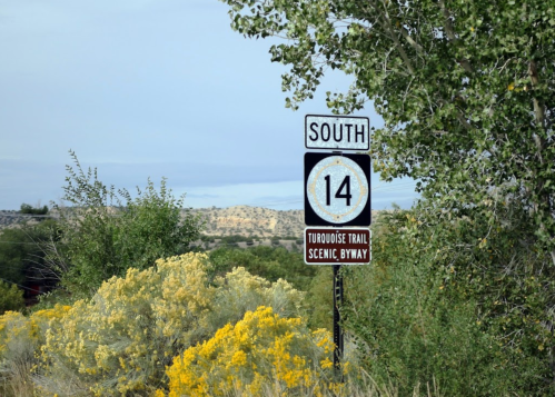 Sign for South 14, Turquoise Trail Scenic Byway, surrounded by greenery and yellow flowers against a cloudy sky.