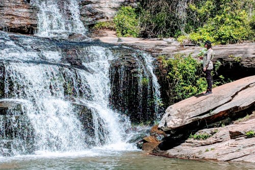 A person stands on a rock by a waterfall, surrounded by lush greenery and flowing water.