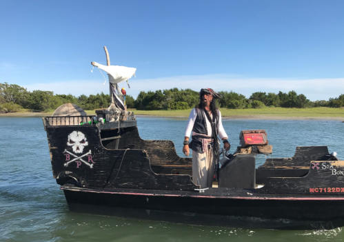 A pirate-themed boat with a man in costume stands on the deck, surrounded by water and greenery under a clear blue sky.
