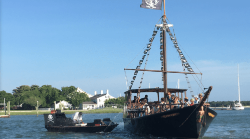 A pirate ship with a black flag sails in a harbor, accompanied by a smaller pirate-themed boat.