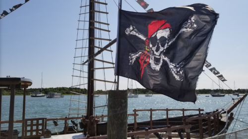 A pirate ship with a skull and crossbones flag sails in a harbor, surrounded by boats and greenery.