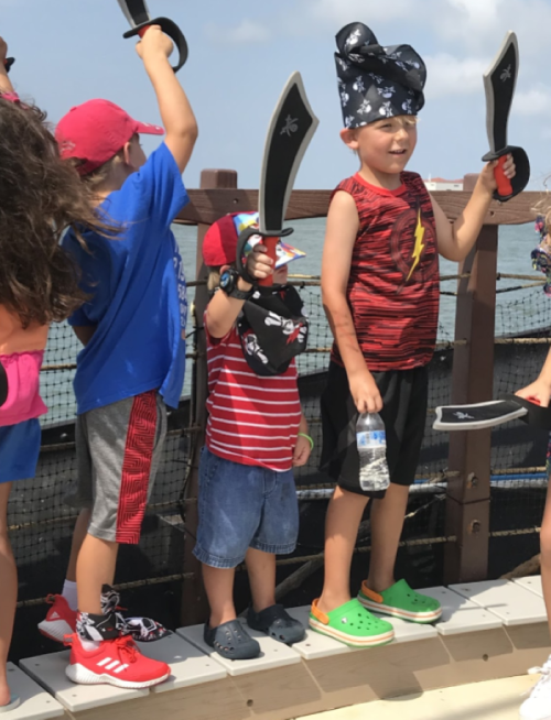 Children dressed as pirates, holding toy swords on a boat, with a sunny sky and water in the background.