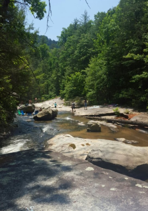 A serene river scene with rocky banks, surrounded by lush green trees and two people walking along the water.