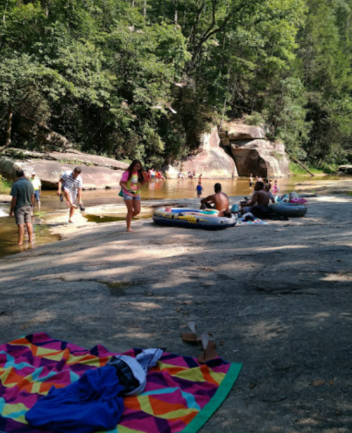 People enjoying a sunny day by a river, with colorful towels and inflatable rafts on a rocky shore surrounded by trees.