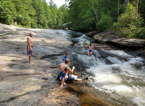 A group of people enjoying a river, with two children playing in the water and adults relaxing on the rocks nearby.