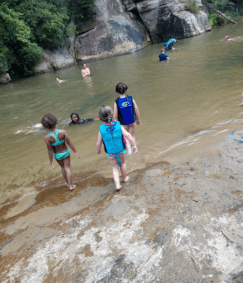 Children in swimsuits play by a river, with some swimming and others wading in shallow water. Green trees in the background.