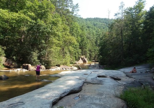 A serene river scene with people wading and relaxing on smooth rocks, surrounded by lush green trees and mountains.