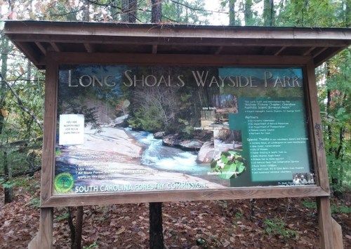 Sign at Long Shoals Wayside Park, featuring park information and a scenic view of a river surrounded by trees.