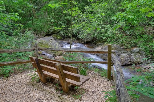 A wooden bench by a serene stream, surrounded by lush green trees and rocky terrain.
