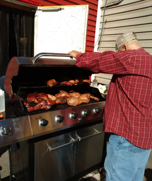 A person in a red plaid shirt grills meat on a large barbecue in a backyard setting.