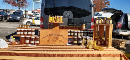A market stall displays jars of honey and beeswax products, with a sign reading "Vdovichenko Bee Farm" in front of a van.
