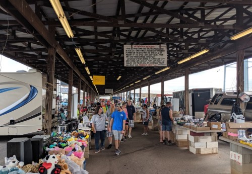 A bustling market scene with people walking between vendor stalls filled with various goods under a covered structure.