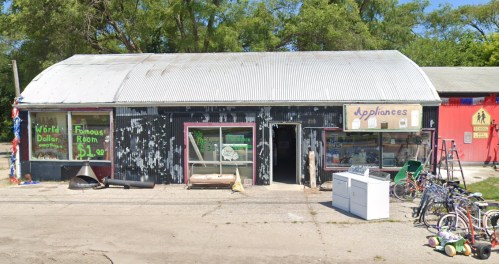 A weathered storefront with signs for appliances and a dollar store, featuring large windows and a metal roof.