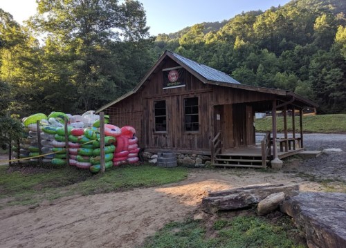 A rustic wooden building surrounded by colorful stacked bags, set in a lush, green mountainous landscape.